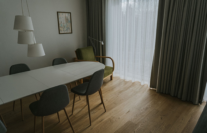 Zielony fotel, biały stół, drewniana podłoga a także szare zasłony wraz z firnami